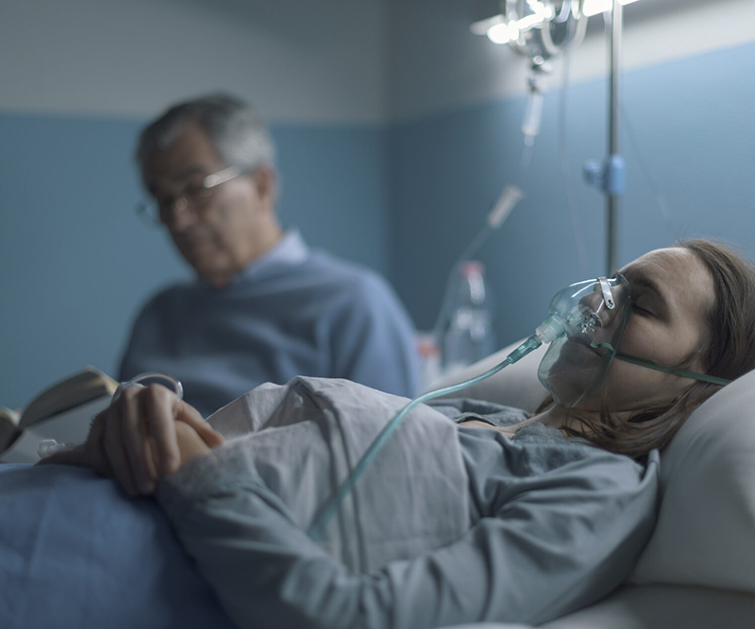 Senior man sitting next to her daughter at the hospital and assisting her at night, she is sleeping and wearing an oxygen mask