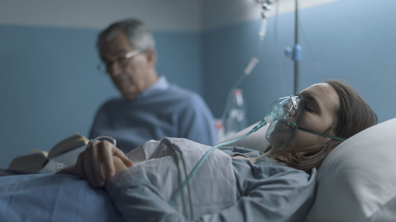 Senior man sitting next to her daughter at the hospital and assisting her at night, she is sleeping and wearing an oxygen mask